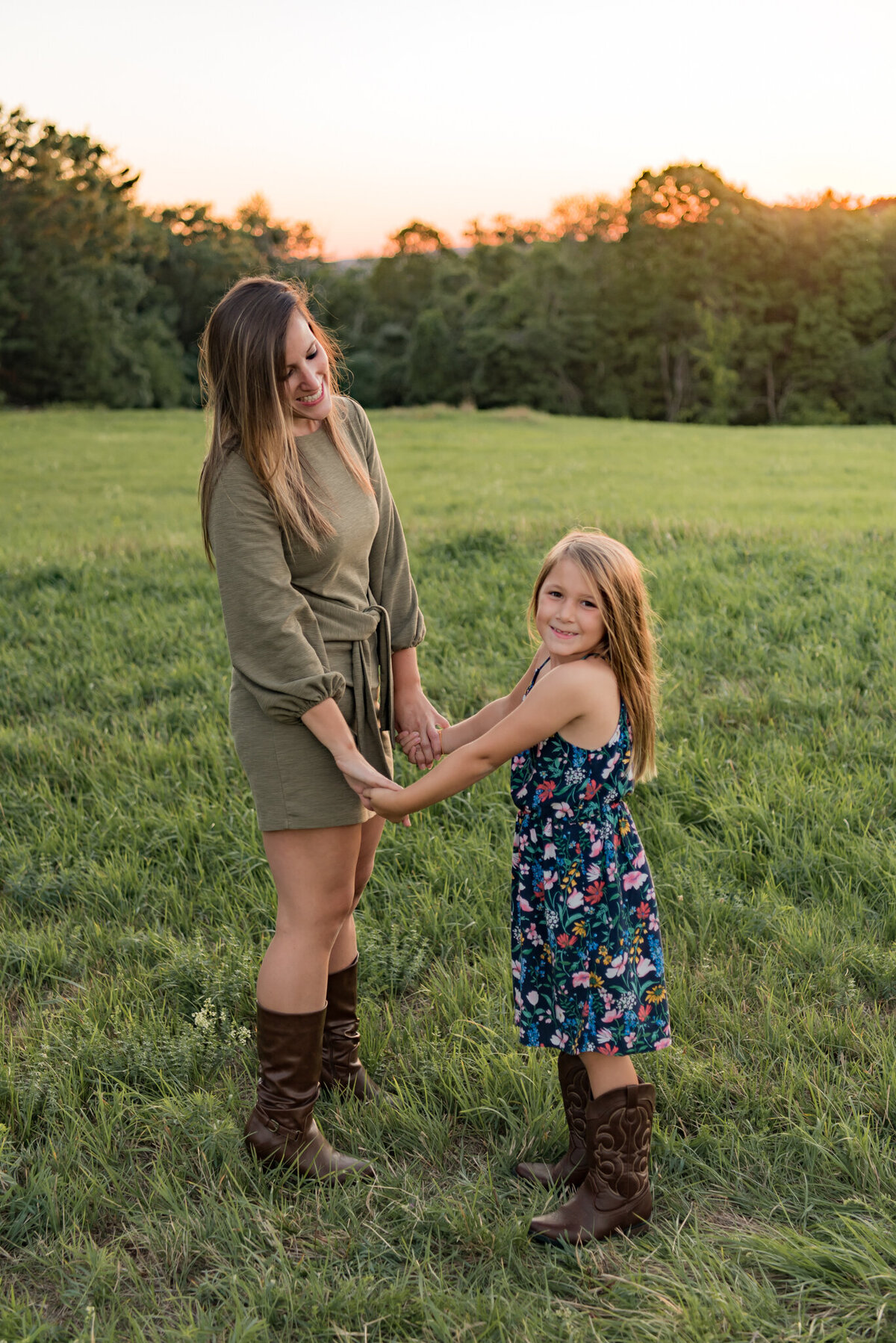 Mother and daughter are holding hands in a green field with the sun setting behind them. The mother is smiling at her daughter while the daughter looks at camera.