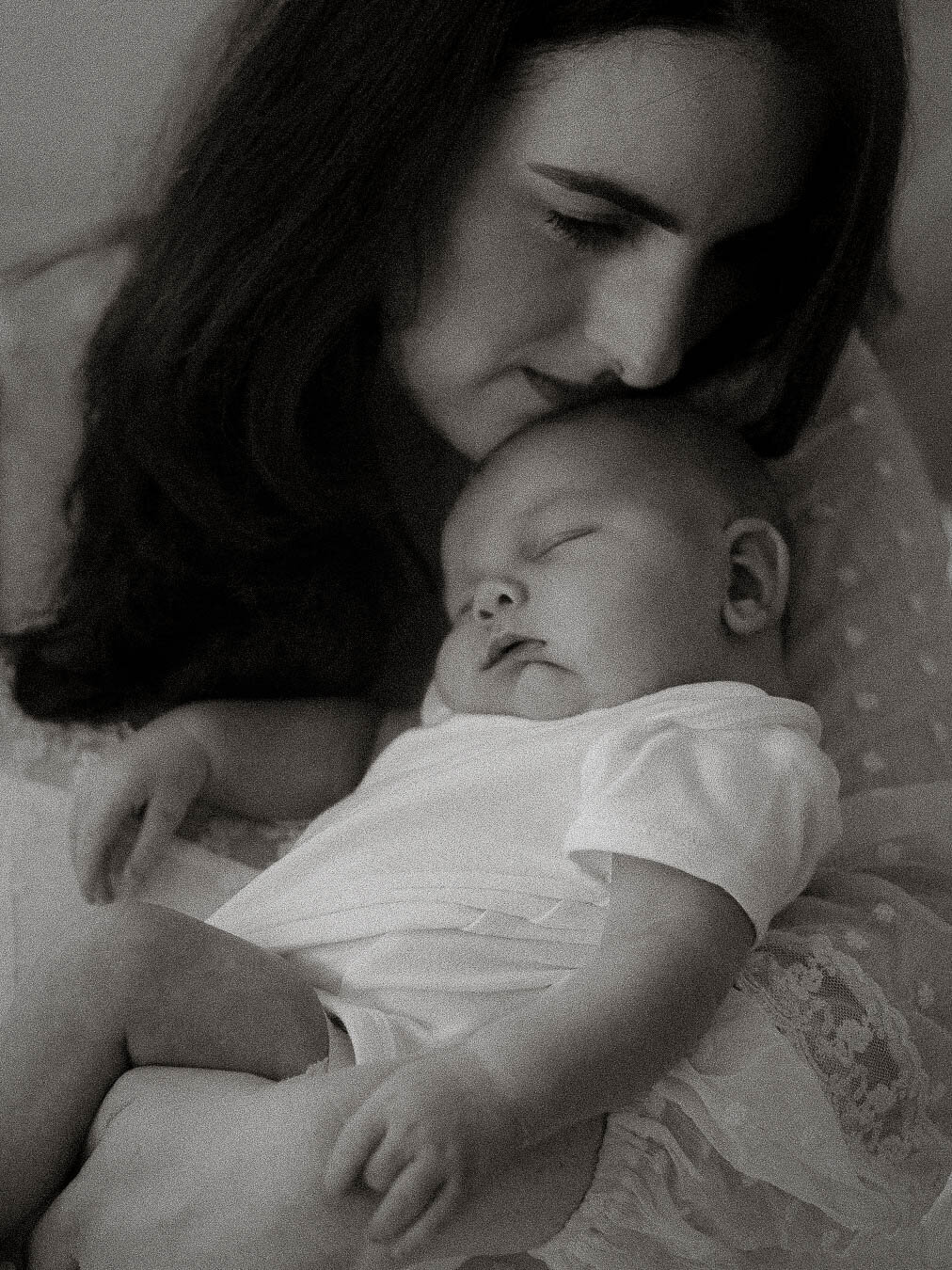 Black and white photo of a mother resting her cheek gently against her baby’s head while the baby sleeps peacefully in her arms.