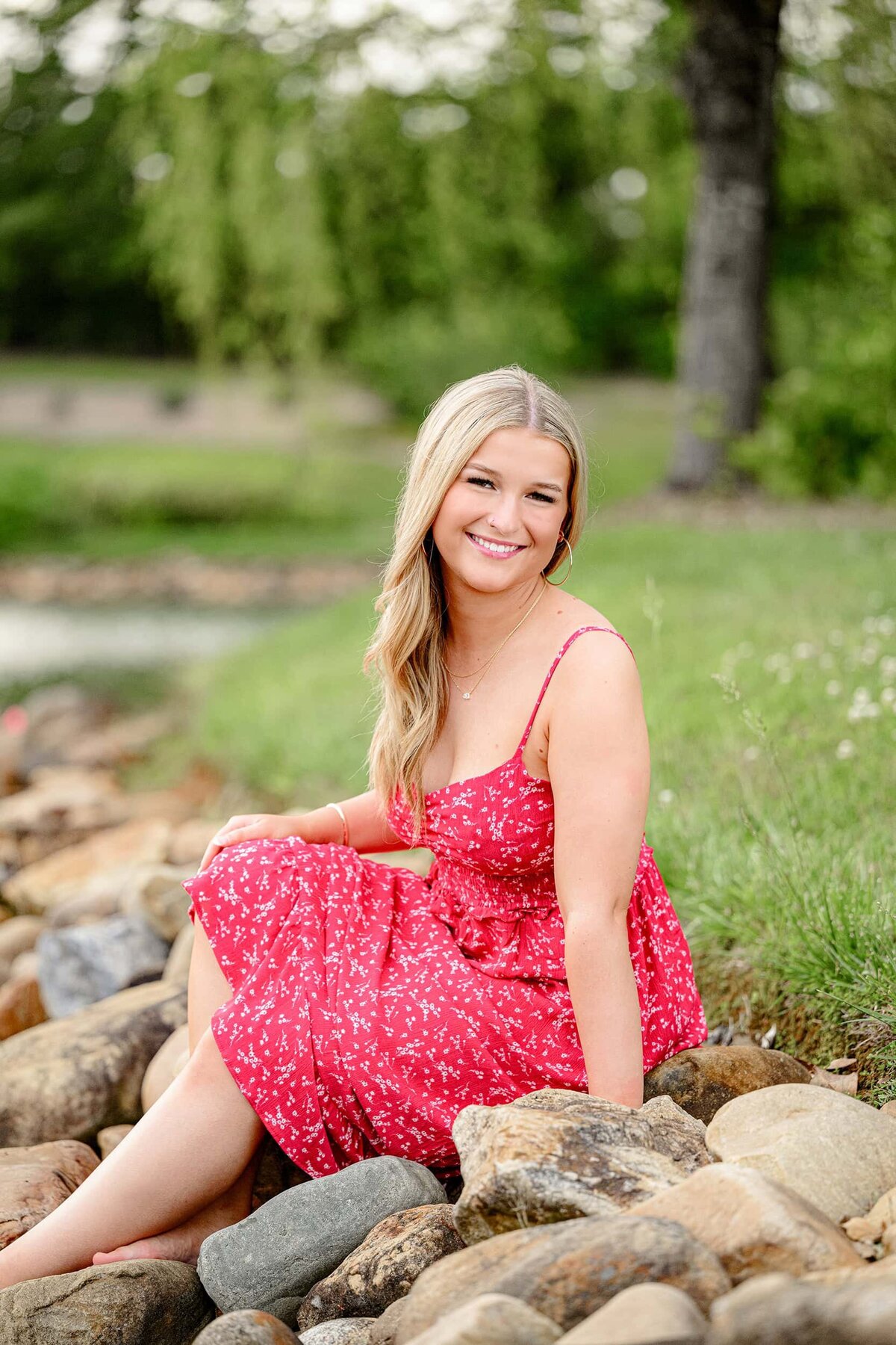 Charlotte senior photoshoot with a blonde graduate wearing a red dress sitting on a rock bank for her portraits