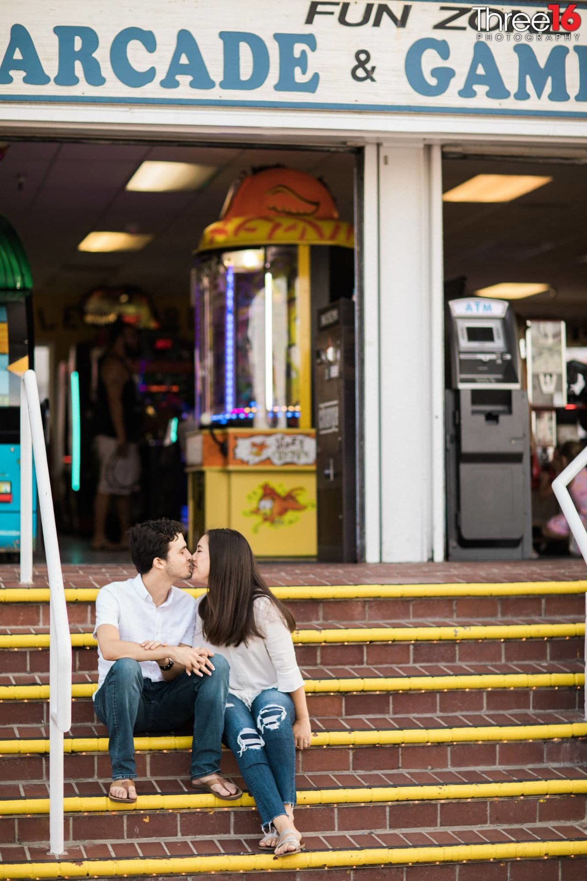Engaged couple share a kiss while sitting on the steps to the Balboa Fun Zone Arcade