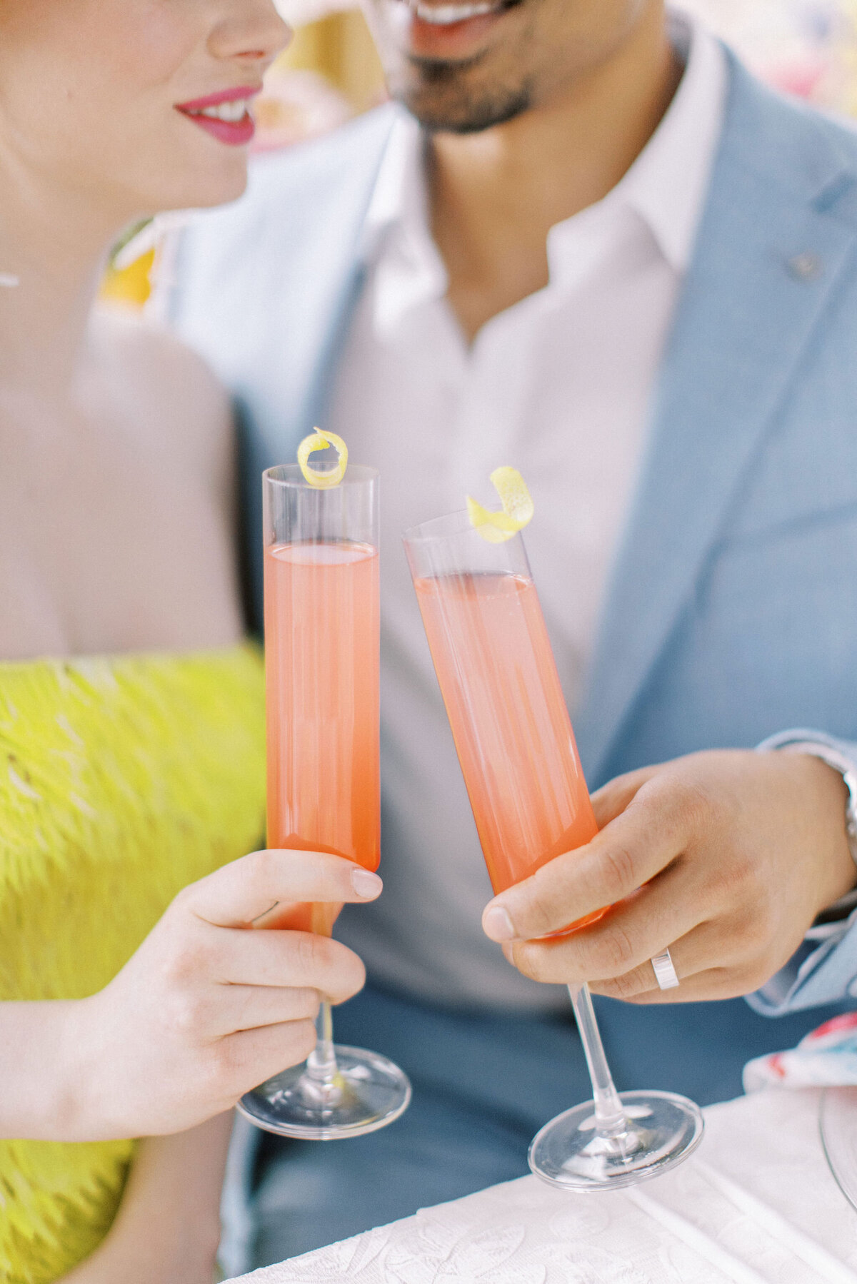 A couple toasts with tall glasses of a pink drink garnished with lemon twists. The woman wears a yellow dress, and the man sports a light blue suit. Only their torsos and hands are visible, capturing the essence of intimate weddings in Banff, Alberta.