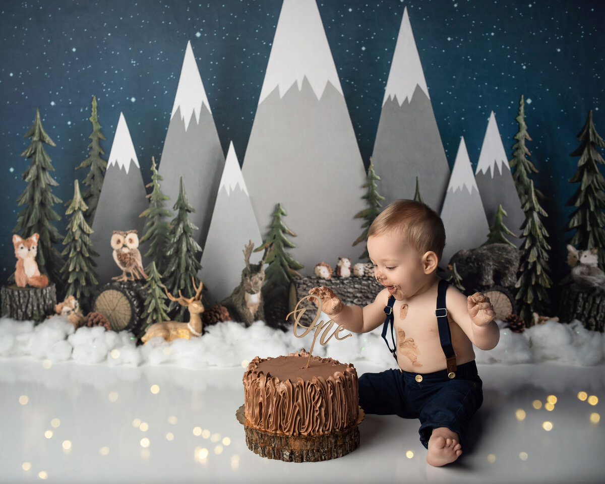 Winter onederland one year old cake smash baby boy wearing denim overalls smiling and looking at his chocolate cake with woodsy winter backdrop with woodland critters and sparkes