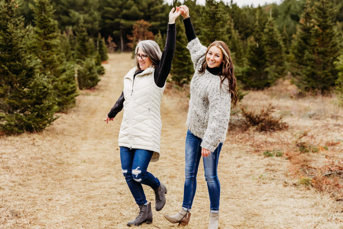 Daughter twirling her mother in a field surrounded by pine trees. Captured by Ashley Kalbus Photography