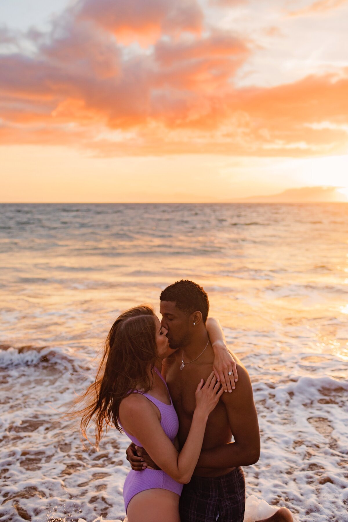 Woman in violet one piece swimsuit kisses her boyfriend while they kneel in the ocean at sunset in Wailea during their engagement session