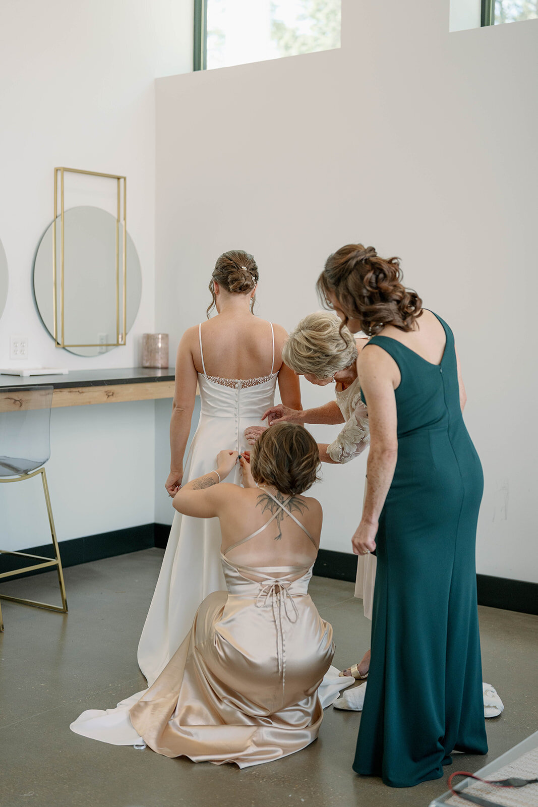 Bride getting ready with her sister, mom, and grandmother helping button up the back of her wedding dress before her ceremony at Leona Road in Grand Rapids MI.