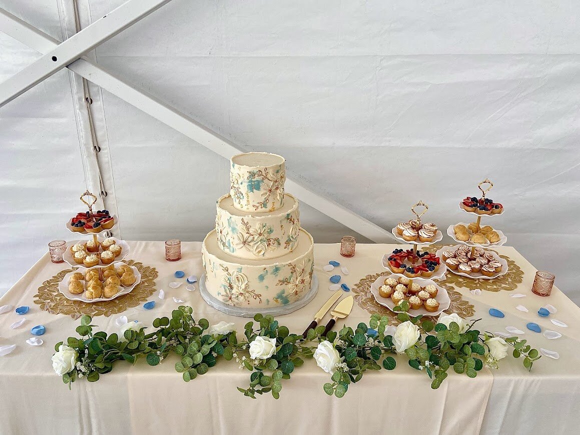 A three tier cake with white frosting, blue accents, and white and gold florals sits on a table surrounded by tiered displays of mini fruit tarts, lemon meringue pies, and cream puffs