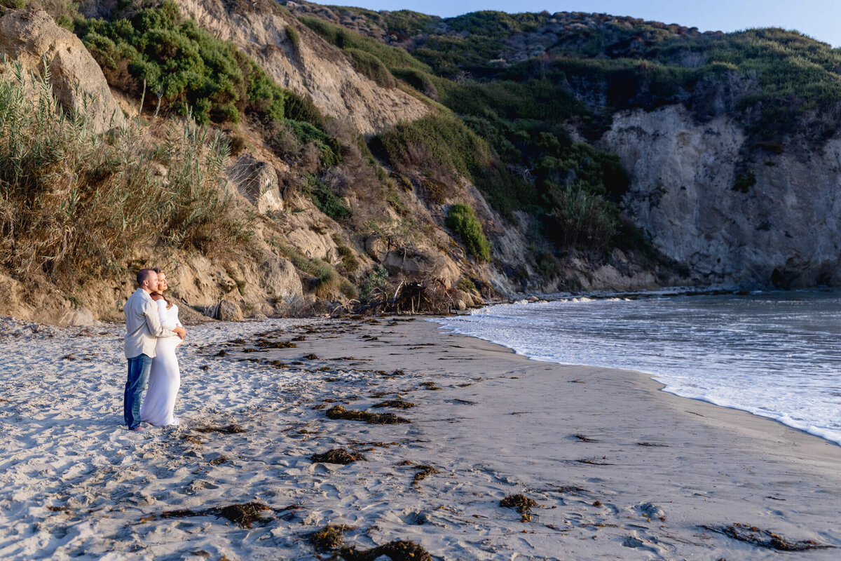 A couple stands on a sandy beach, embracing and looking out at the calm ocean. Cliffs covered with greenery rise in the background under a clear blue sky. Seaweed is scattered across the shoreline.