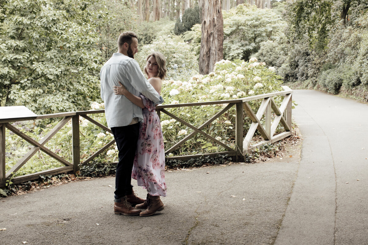 Bride and Groom standing on a mountain top. The wind is blowing in her hair and they are looking out to the view while hugging each other. The bride is a long sleeved wedding gown.