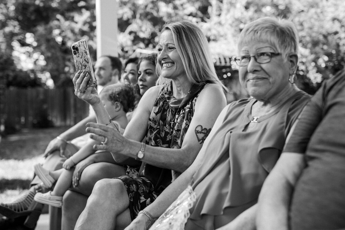 Black and white photo of wedding guests seated during the ceremony, one woman smiling as she takes a picture with her phone