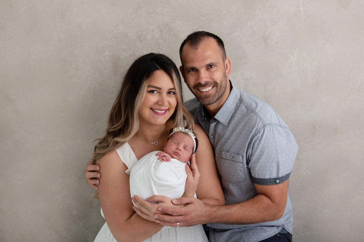 mom and dad holding newborn baby girl on a beige background for their newborn photography session in Burlington, Ontario