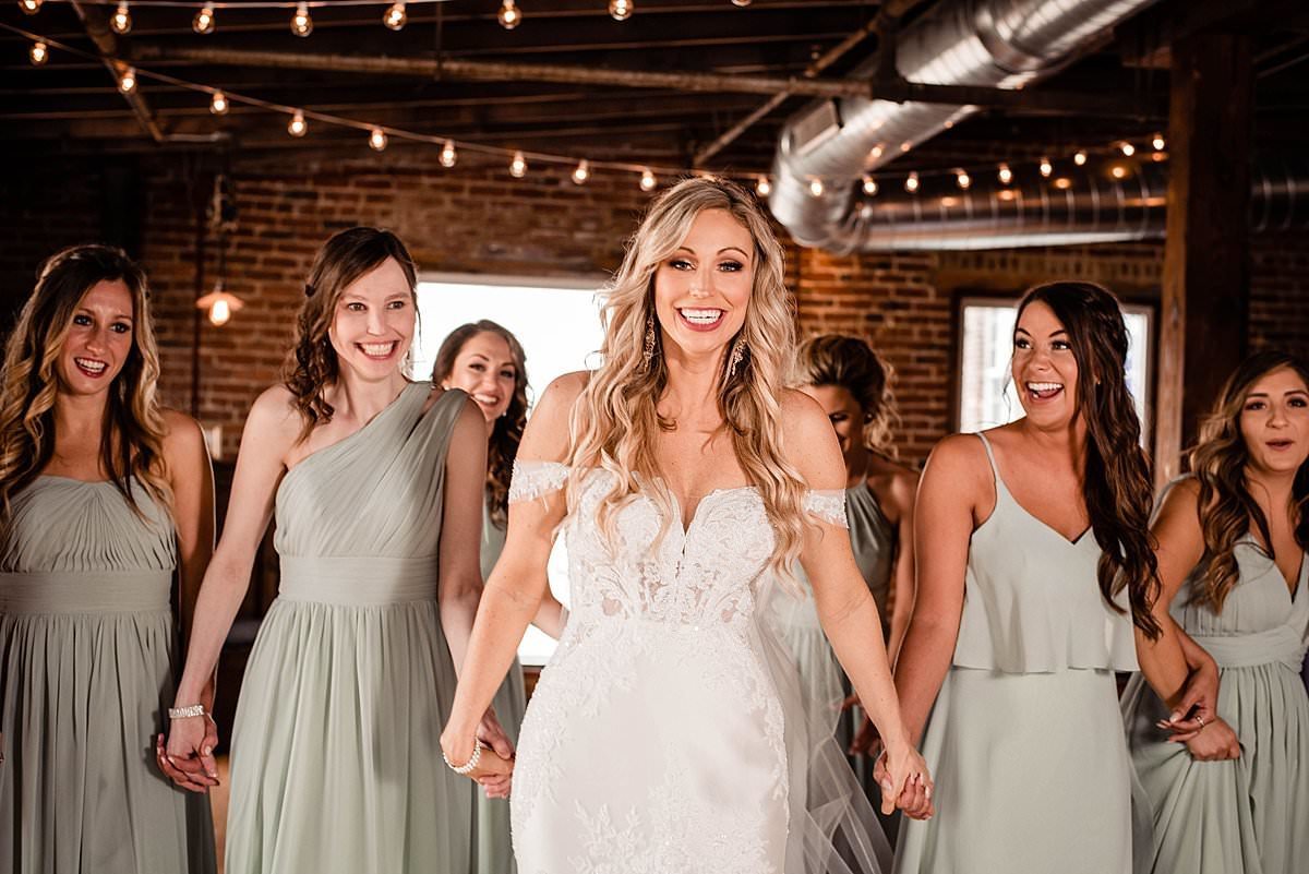 Bride smiling joyfully at camera while holding her friends hands, bistro lights and brick walls surround them