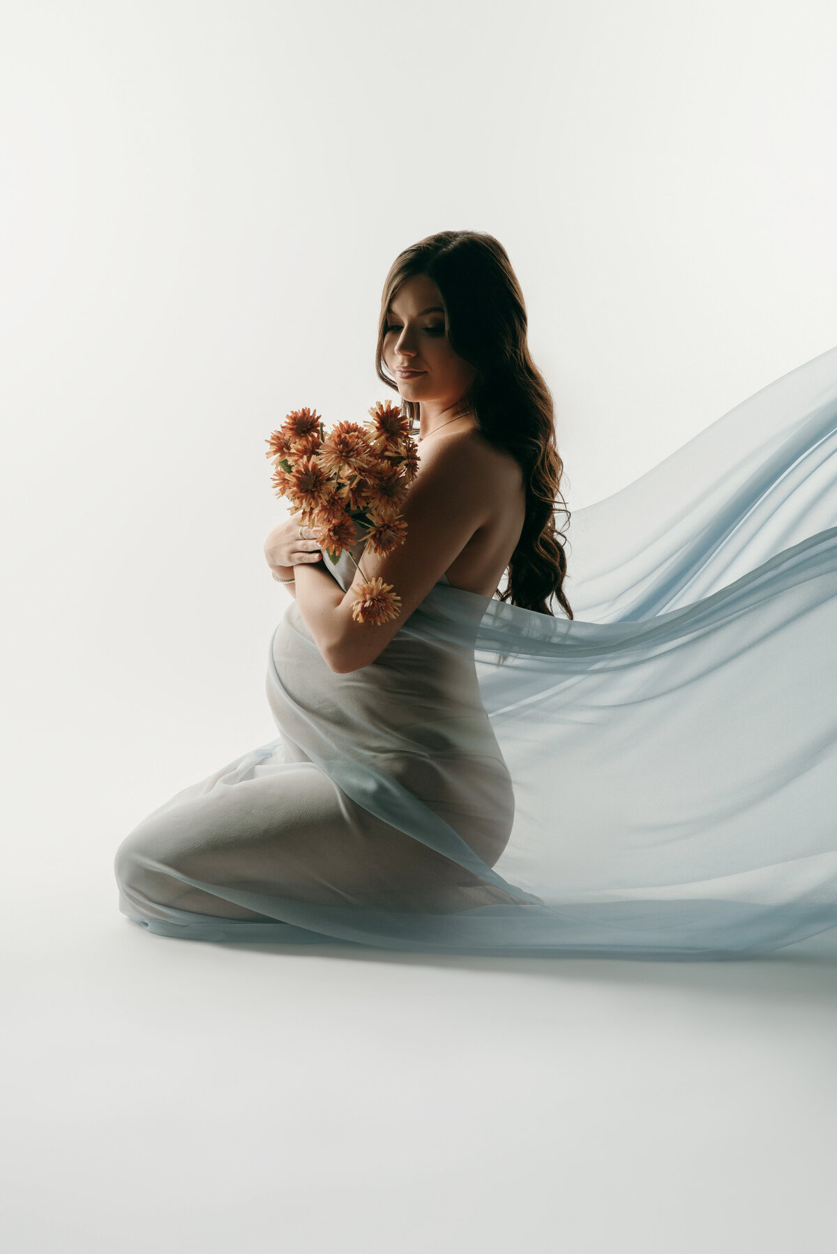 kneeling pregnant woman draped in flowing  baby blue fabric holding flowers over baby bump