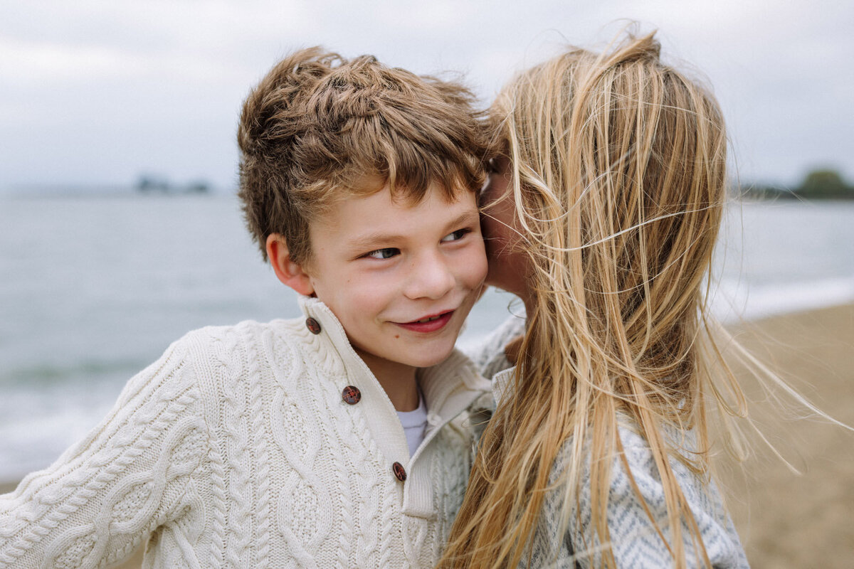 brother leaning over towards sister as she whispers a secret with wind blowing their hair at alameda beach
