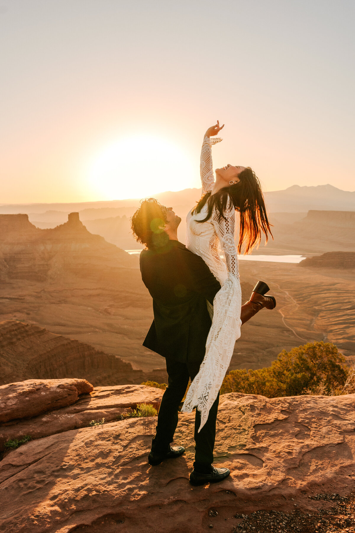 Bride and groom elopement at Moab during sunrise while groom picks up the bride in her Claire Pettibone gown reaching up to the sun