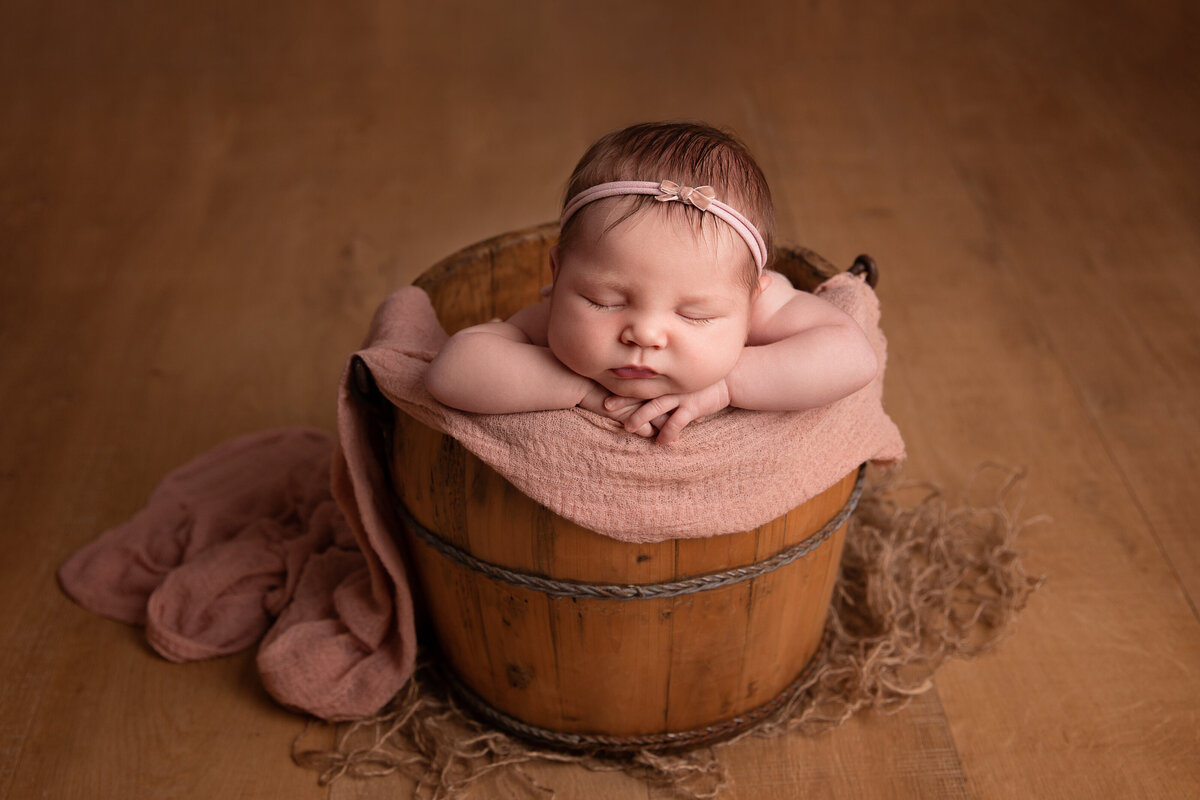 Newborn posed sleeping on her hands in a wooden bucket with a pink wrap draped to the side.