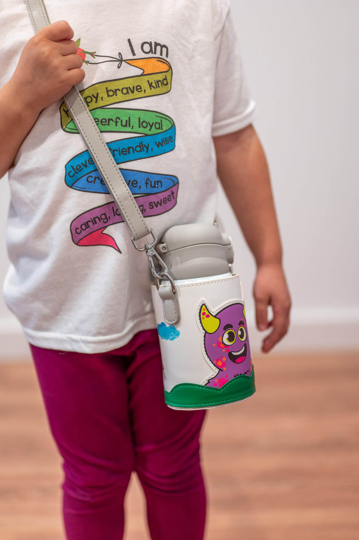 Close up of a girl wearing a white tshirt with words "I am happy, brave, kind, powerful, loyal, fun, friendly, caring" while wearing  a crossbody strap holding a kid's water bottle for commercial brand Luua, located in Wilton, CT.