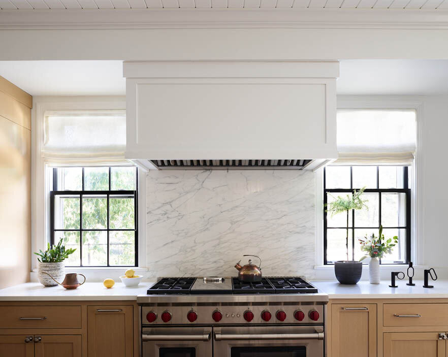 Admire the sophisticated kitchen in a historic Hingham home, featuring a custom hood integrated with the soffit, a slab marble backsplash, and a perfectly positioned range between original windows.