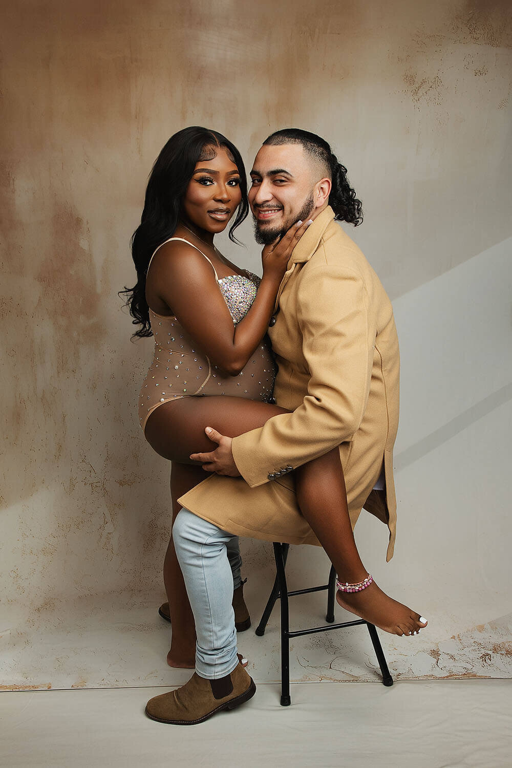 A man in a tan overcoat sits on a stool in a New Orleans Maternity Photographer studio with his pregnant wife putting one leg up on him in a silver one-piece maternity suit