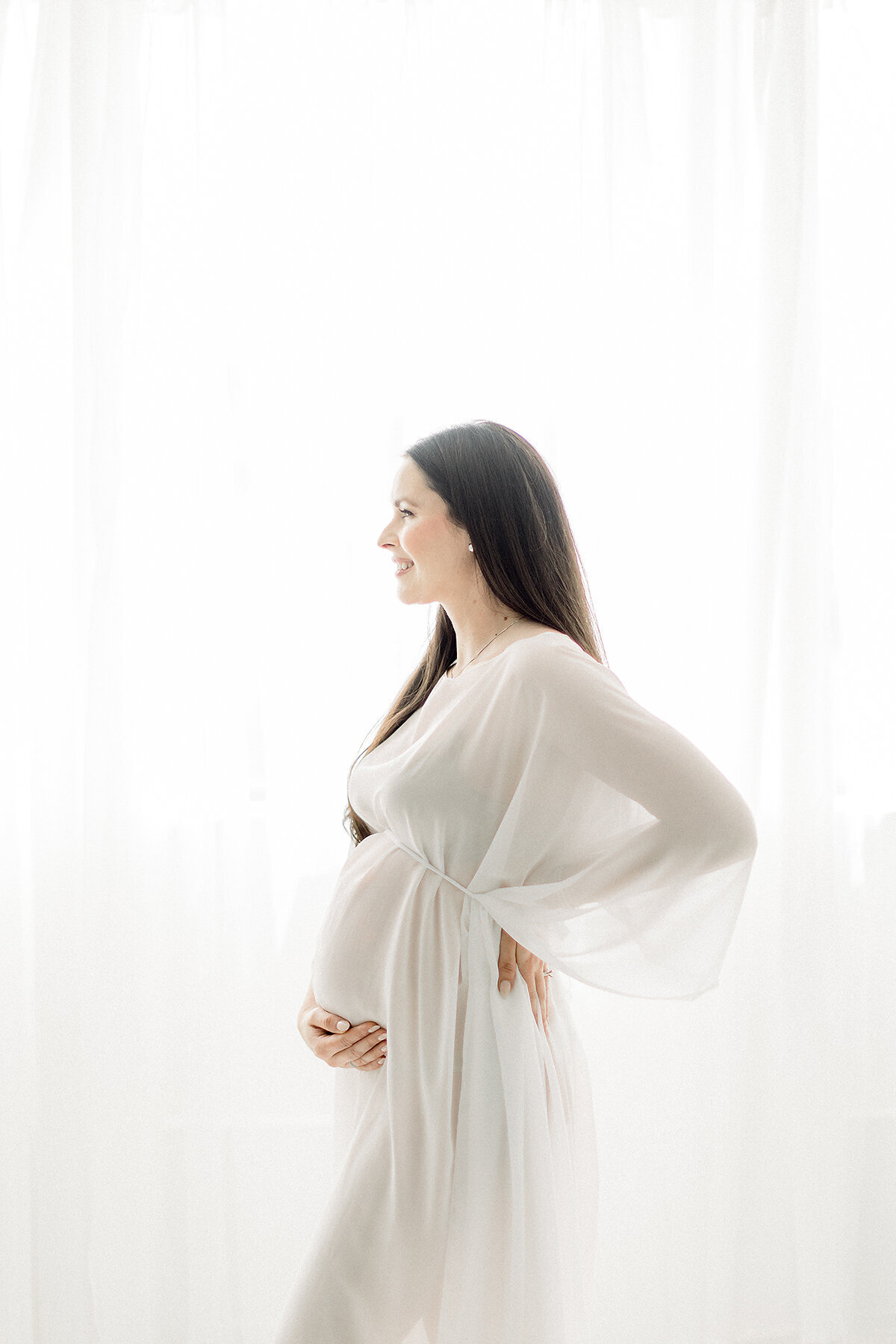 A pregnant mother dressed in a ivory chiffon maternity gown while she is standing in front of a window embracing her growing belly at a Dallas/Fort Worth photography studio.