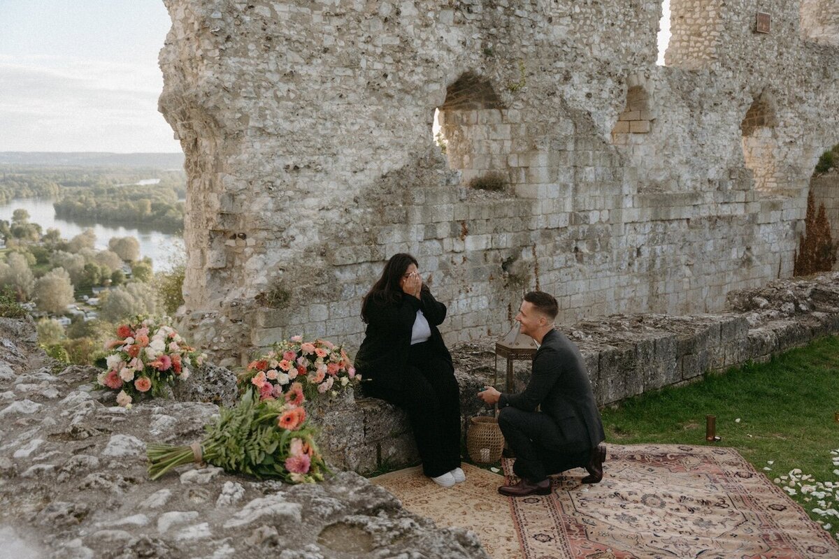 Homme agenouillé faisant sa demande en mariage à sa compagne émue. Décor de de ruines médiévales, compositions florales et tapis persan. Par Kamescop, vidéaste et photographe en bretagne.