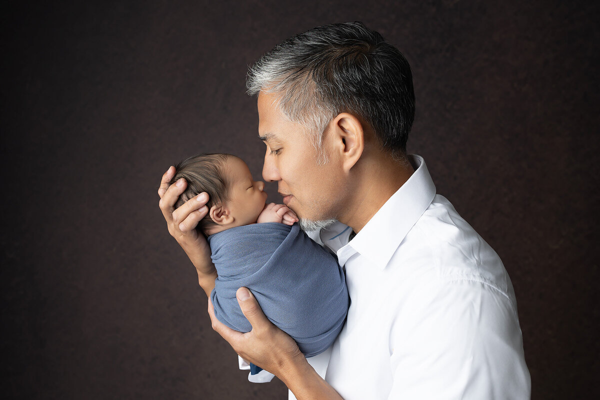 A loving father gently touches noses with his newborn baby wrapped in blue, both sharing a quiet and tender moment.