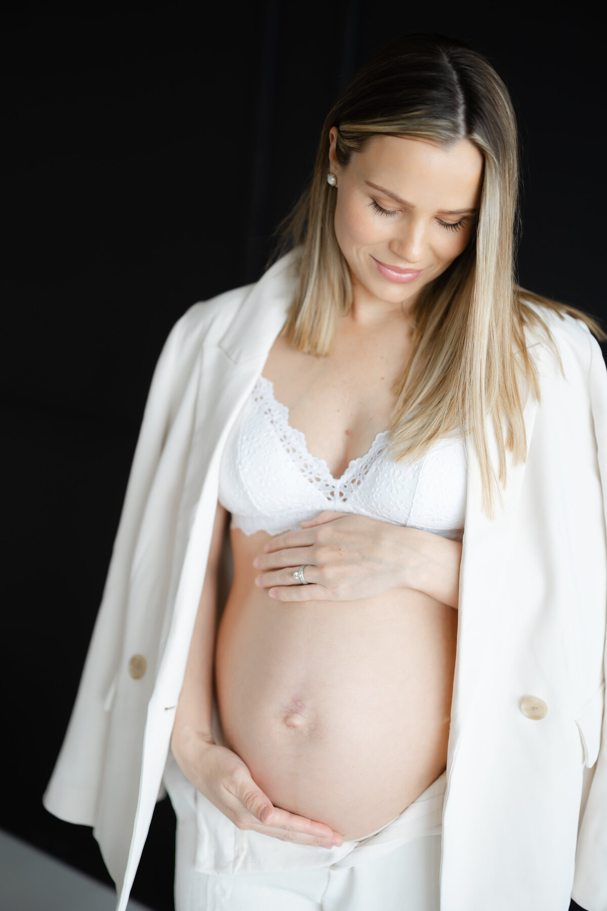 a mother stands in a white pantsuit while holding her pregnant belly as she stands in front of a black wall