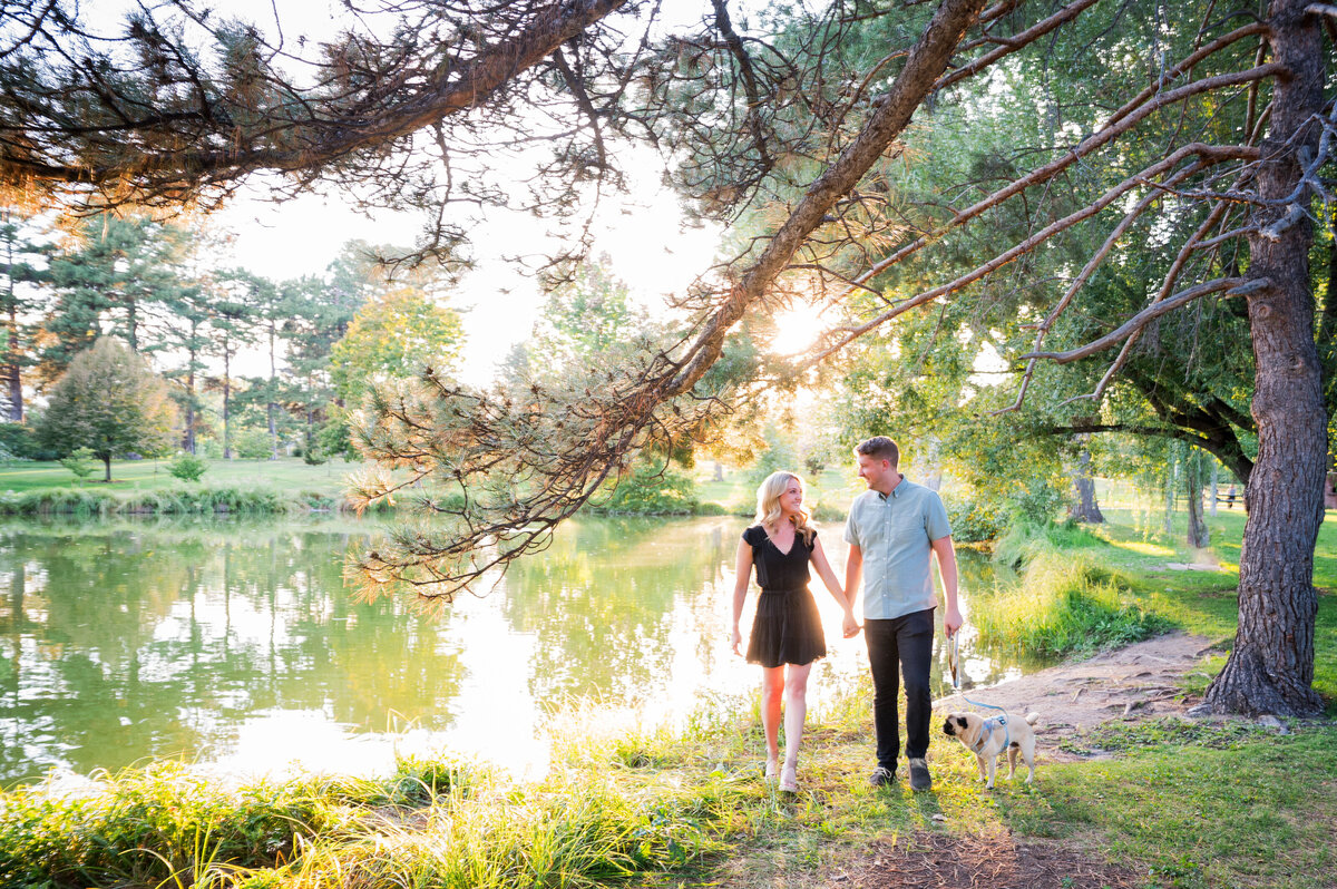 A man and woman walk toward the camera holding hands with a pond and trees in the background.