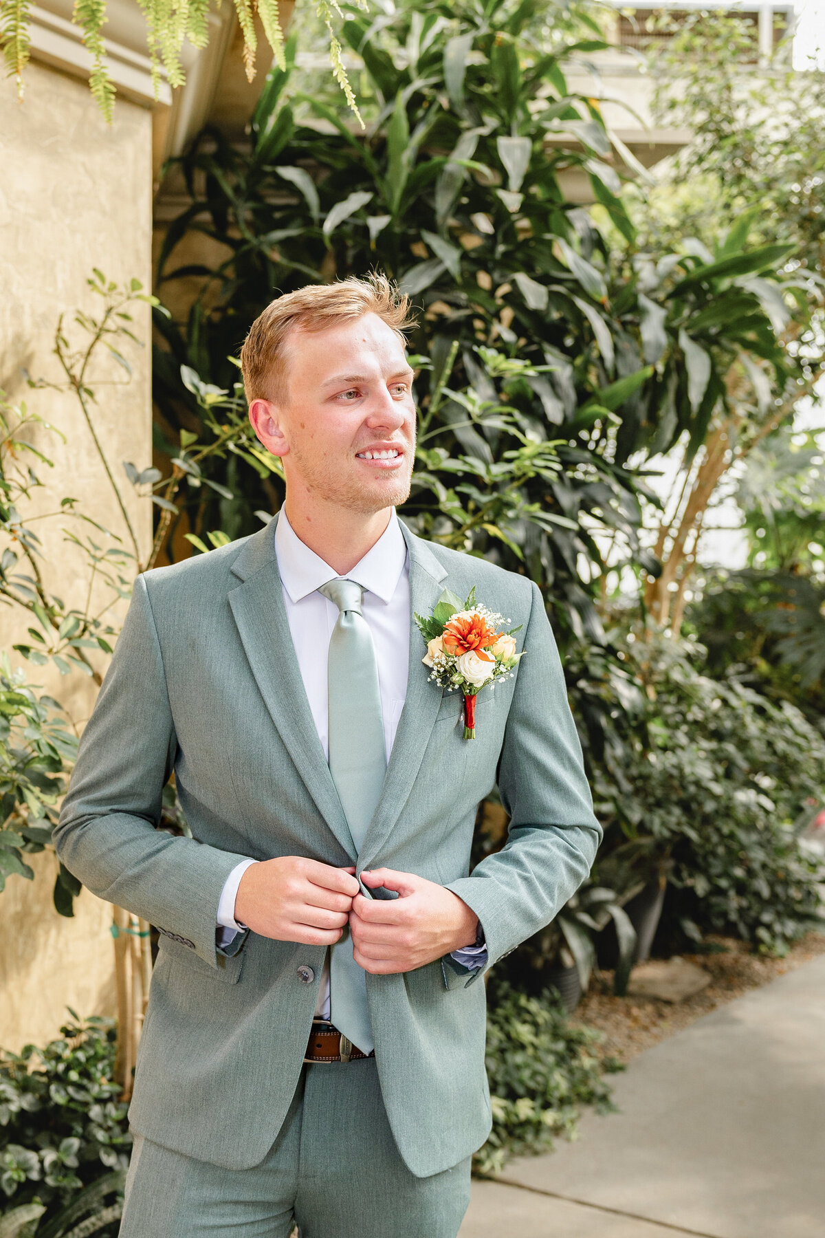 A groom in a sage green suit stands confidently in front of lush tropical plants, adjusting his suit jacket. His boutonniere, made of orange and white flowers, adds a pop of color.