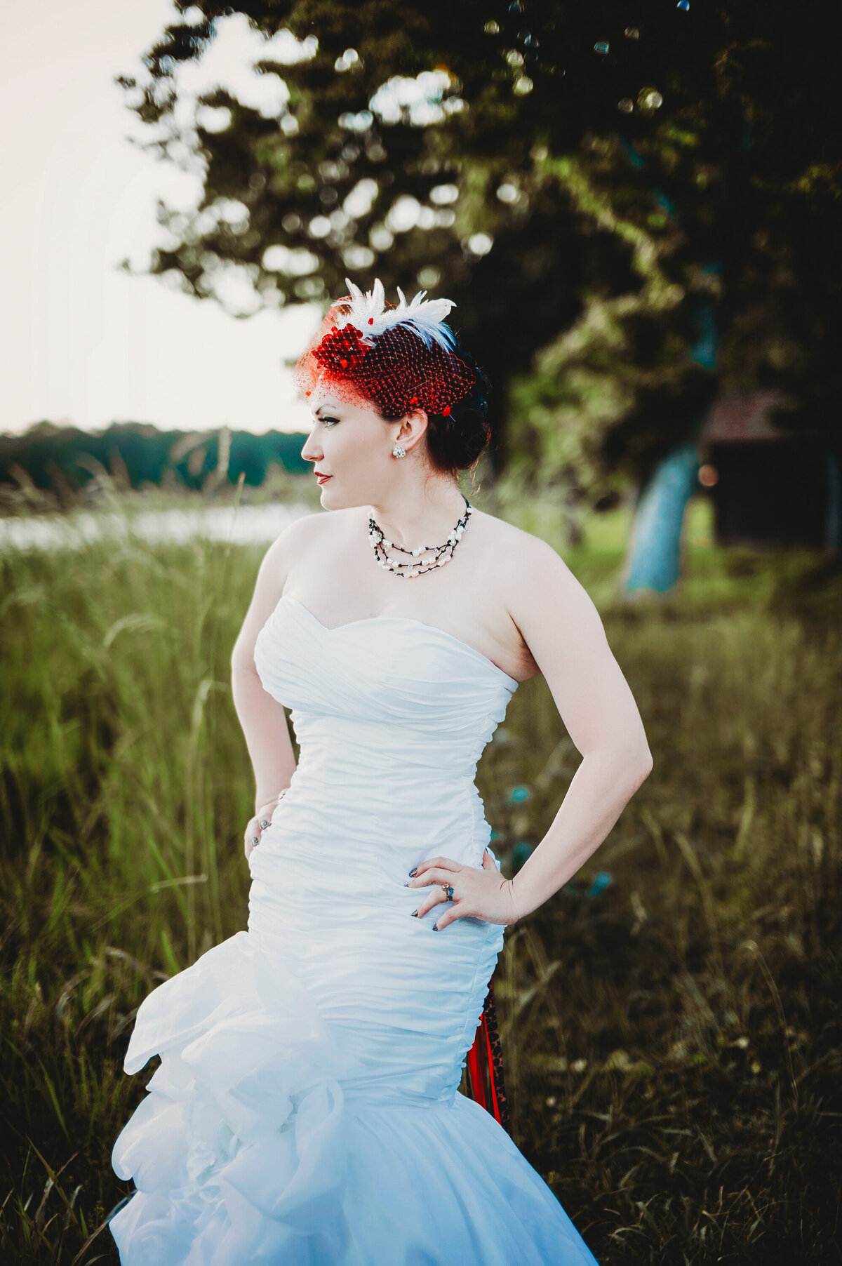 Bride posing in a white wedding dress with a red feather headpiece during an outdoor bridal portrait session in a grassy field in Central Florida.