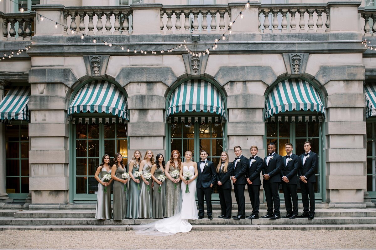 A large groom of elegantly dressed men and women stand together for a portrait outside at the Anderson House.