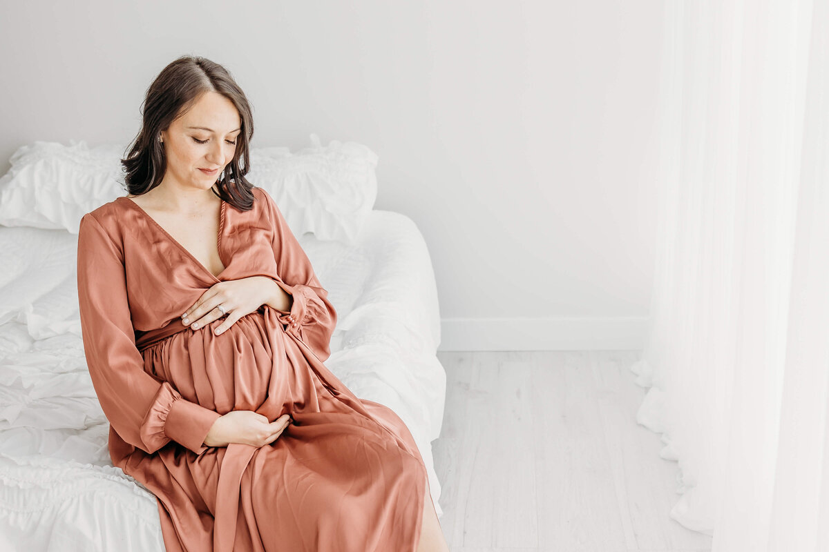 Mom looking down while holding pregnant belly and sitting on  bed in dark pink dress