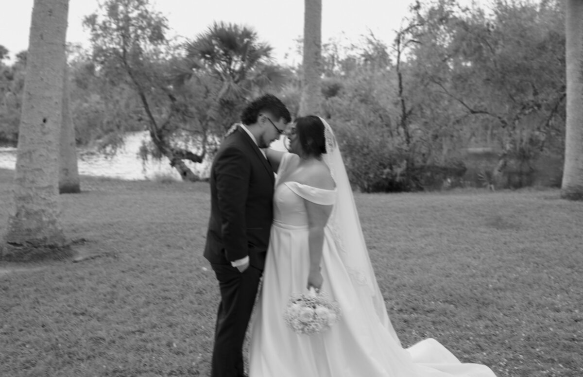 A black-and-white photograph of a bride and groom standing outdoors, gazing at each other