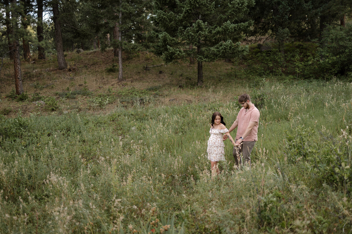 expectant mom leading expectant dad with hands held walking in meadow during lifestyle maternity session in boulder colorado with alexis adkins photography
