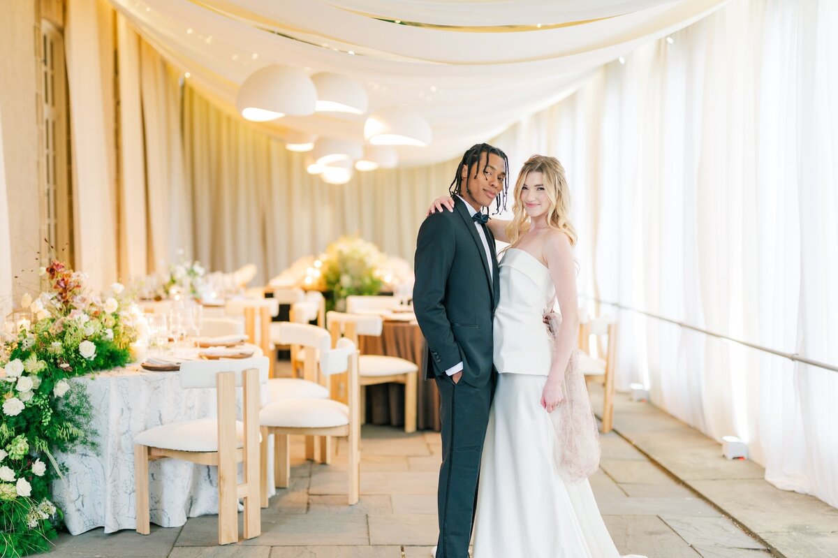 A bride leans against her groom with her arm around his neck. 