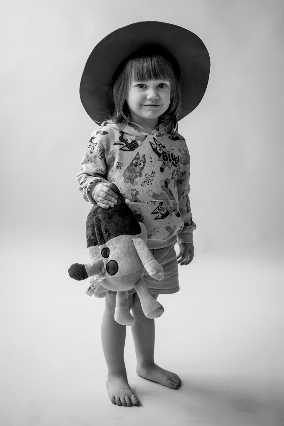 Black and white photo of girl wearing a Bluey sweater and dark colored hat smiling at camera holding a stuffed animal Bluey.
