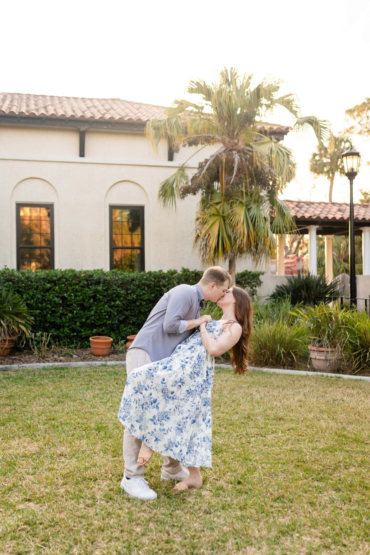 A couple shares a playful and romantic dip kiss in front of Mediterranean-style architecture during their engagement session in Jacksonville, Florida. The bride-to-be wears a flowing blue and white floral dress, while her fiancé supports her in a relaxed button-down and light pants. Surrounded by palm trees, soft golden hour light, and lush greenery, the moment is full of charm and joy. Captured by Heartful Films, a Jacksonville engagement photographer and videographer specializing in stylish, heartfelt storytelling.