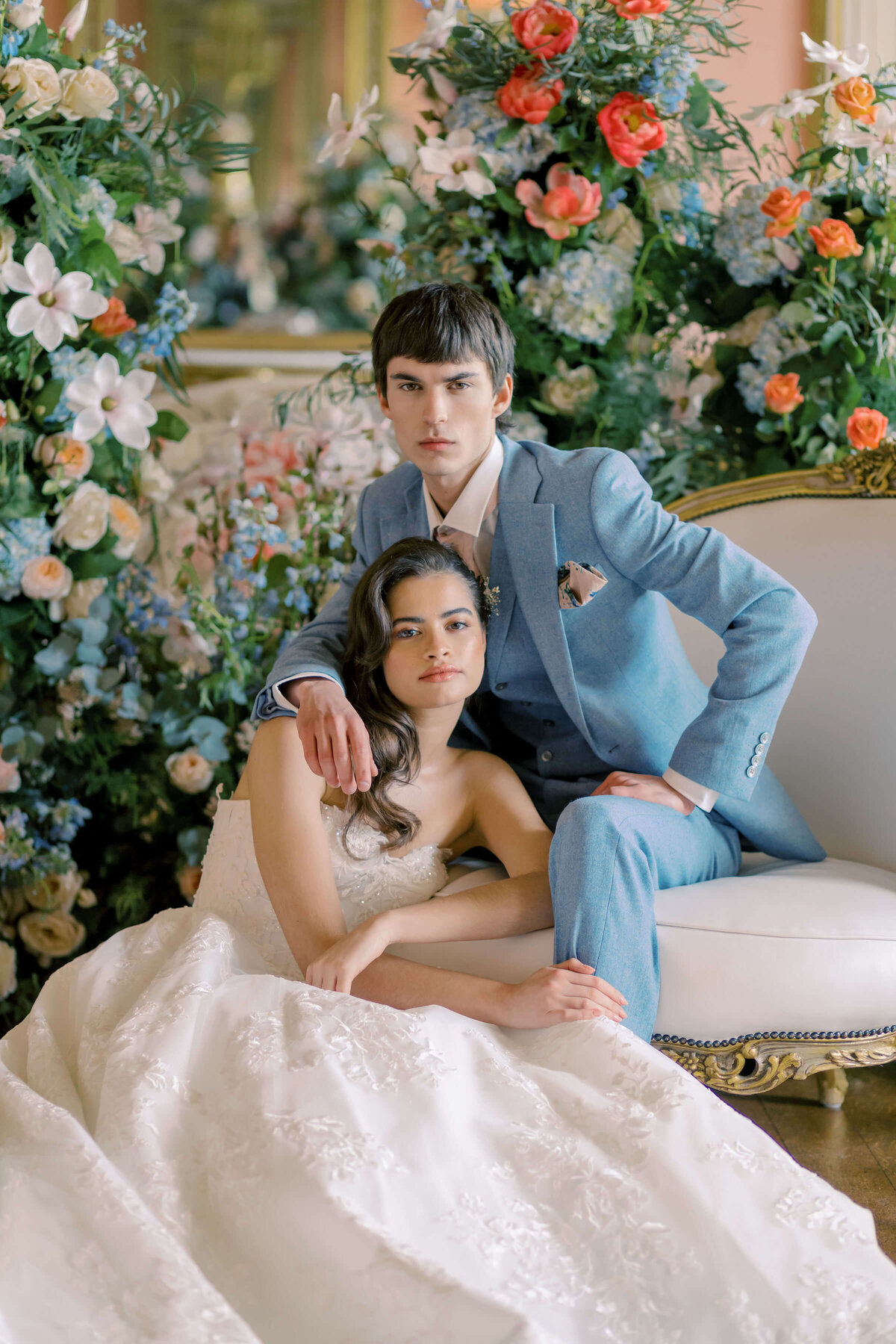 bride sitting on the floor leaning against the groom who is wearing a  blue suit and sitting on a white and gold chair their backdrop is a blue and coral flower wall