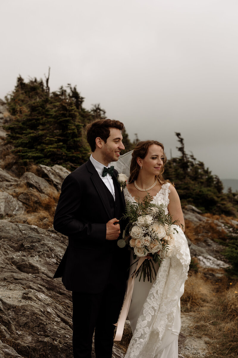 A bride and groom stand together outdoors, surrounded by rocky terrain and evergreen trees, in a scene perfect for Elopement Photography New England. The bride holds a bouquet of white flowers, both are smiling elegantly, with the bride in a white gown and the groom in a black suit.