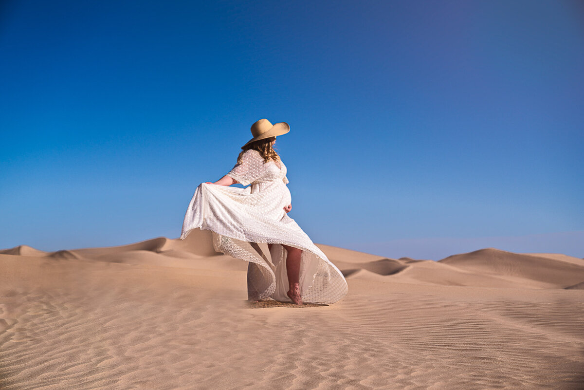 Expectant mom in long white dress and wide brim hat is standing in the desert with her gown blowing behind her. She is standing side profile to the camera. Captured by Bryn Mwar Philadelphia and Main Line photographer Liza Nicole.