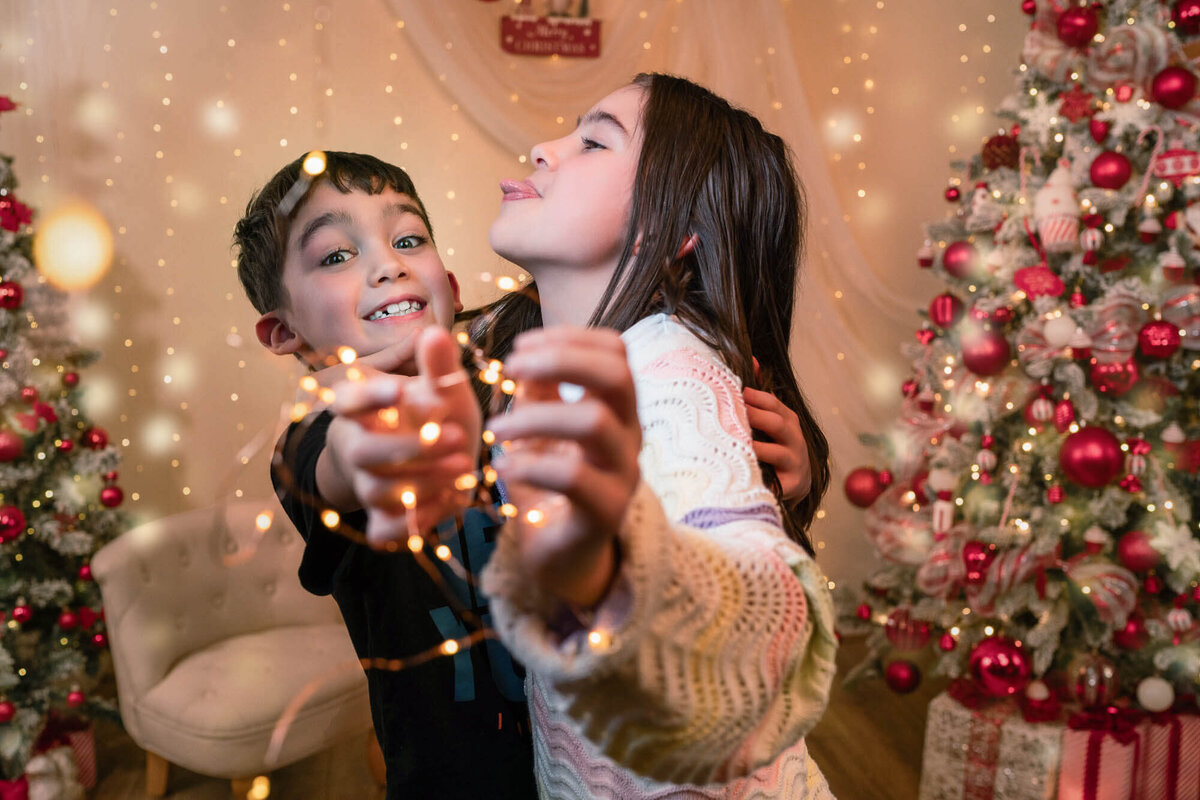 Brother and sister holding Christmas fairy lights in Calini Weddings Studio - festive moment by the tree with warm golden tones.