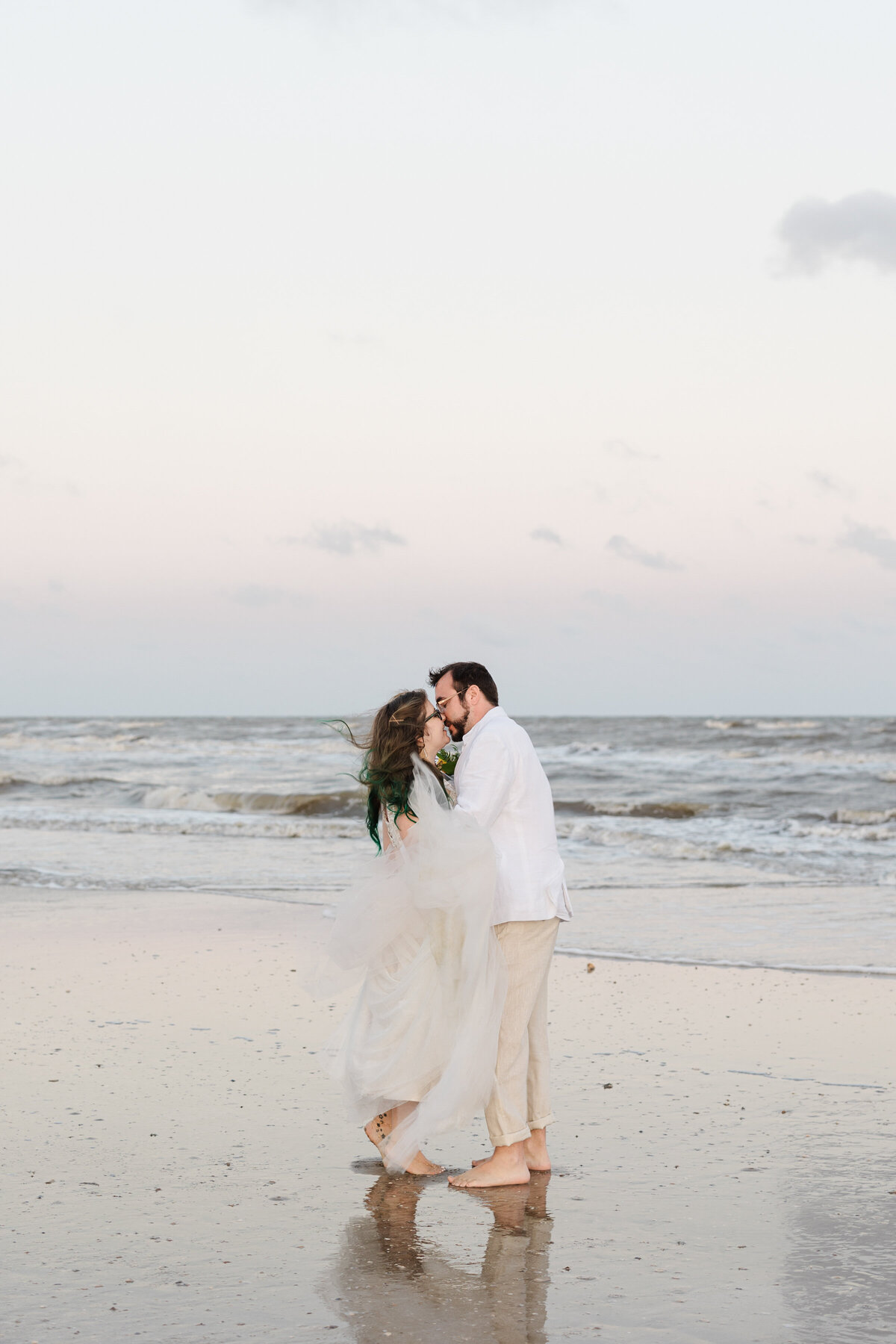 A portrait of a bride and groom sharing a kiss on the beach after their wedding ceremony at Crystal Beach, Texas. The bride is on the left and is wearing a white dress with a long, flowing veil. The groom is on the right and is wearing a white dress shirt with khaki pants.