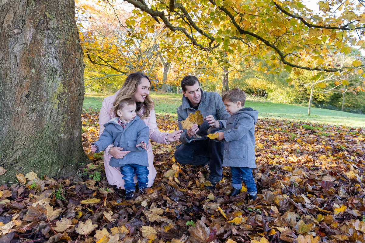 A family of four, including two young children, enjoys collecting autumn leaves in a park filled with colorful fall foliage.