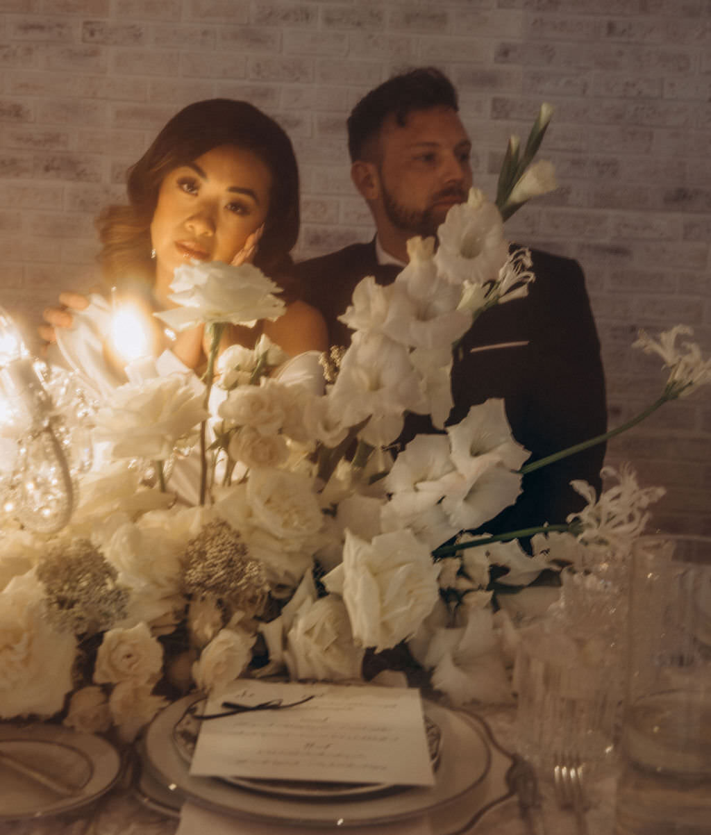 A newlywed couple sitting at a reception table with flowers in front of them.