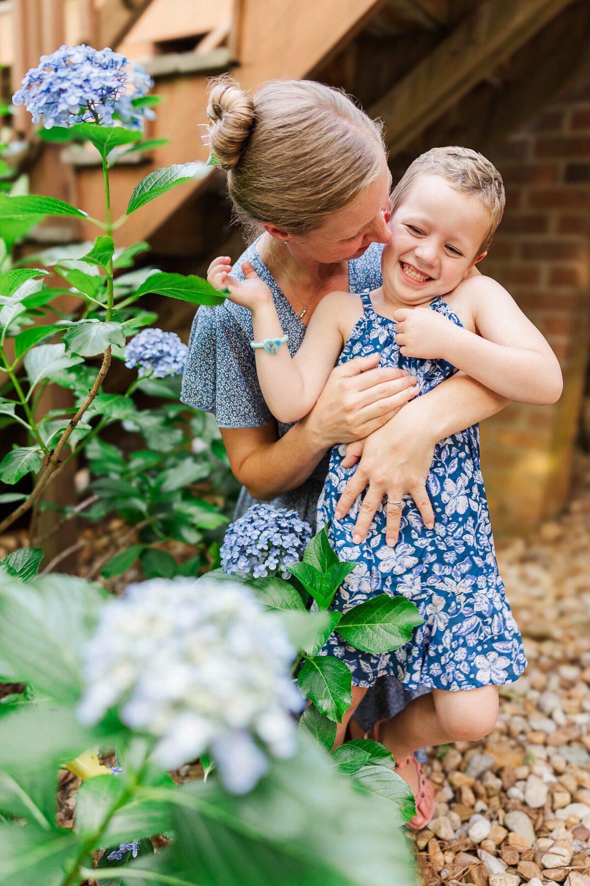 A Fayetteville NC family photographer captures a woman with tied-back hair embracing a laughing child. Both wear blue floral-patterned dresses, surrounded by green leaves and blue hydrangeas against a brick wall backdrop.