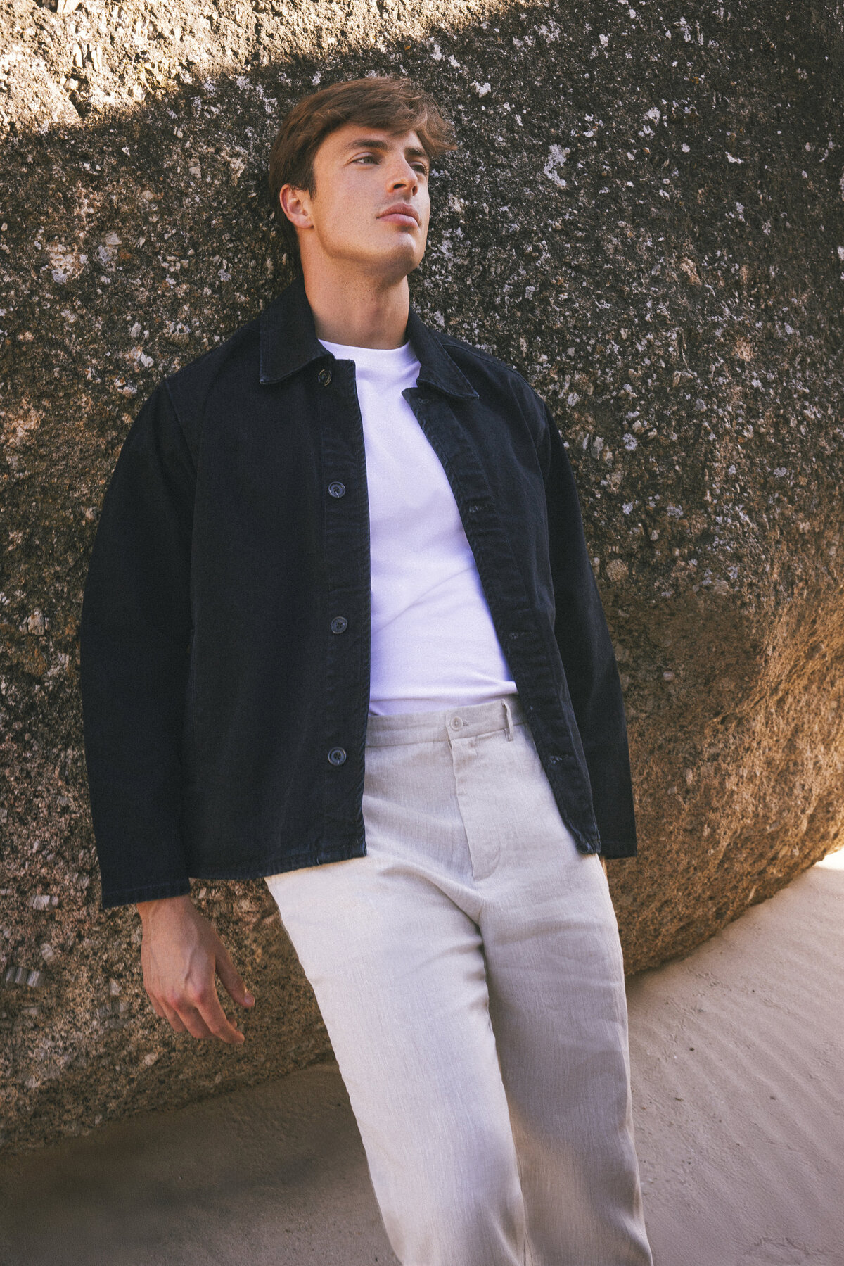 Male model leaning against a large rock in Cape Town, wearing a black denim jacket, white T-shirt, and beige trousers, with soft sunlight casting shadows on the sand.