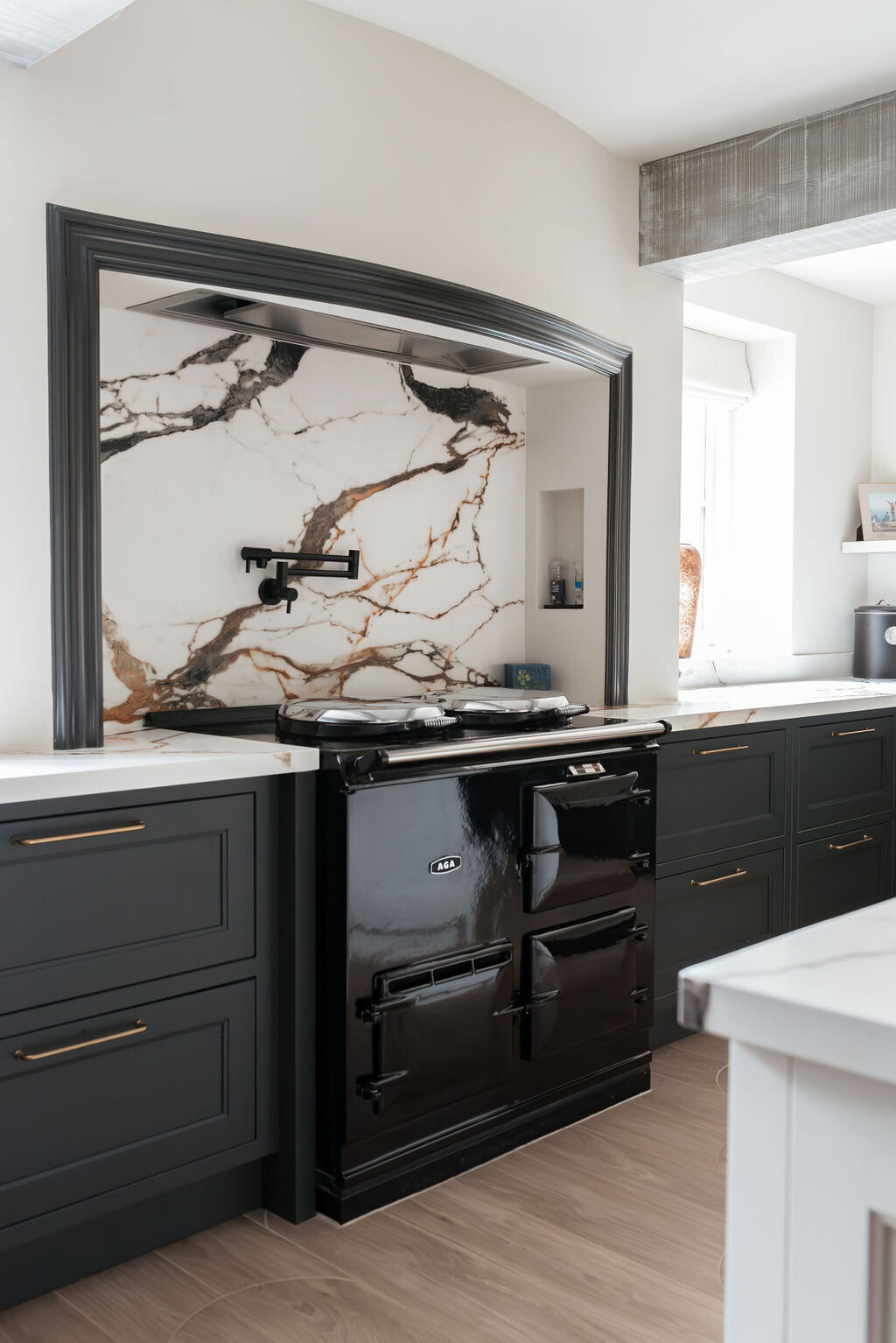 A modern kitchen featuring dark lower cabinets with a white marble countertop. A sleek black stove has a matching marble backsplash and black potfiller.