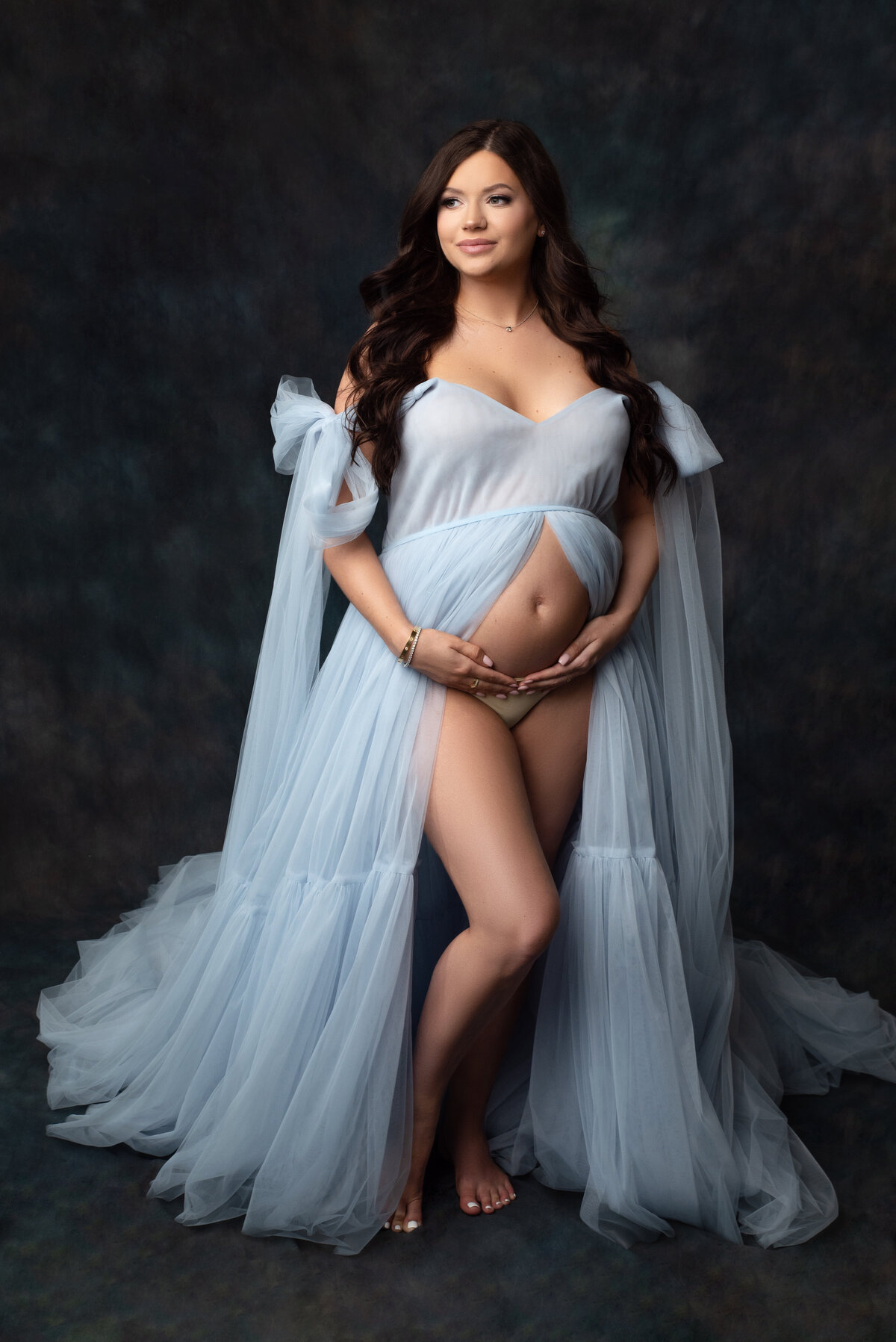 pregnant woman with long brown hair wearing baby blue flowing gown with exposed baby bump looking into the distance on dark green canvas backdrop