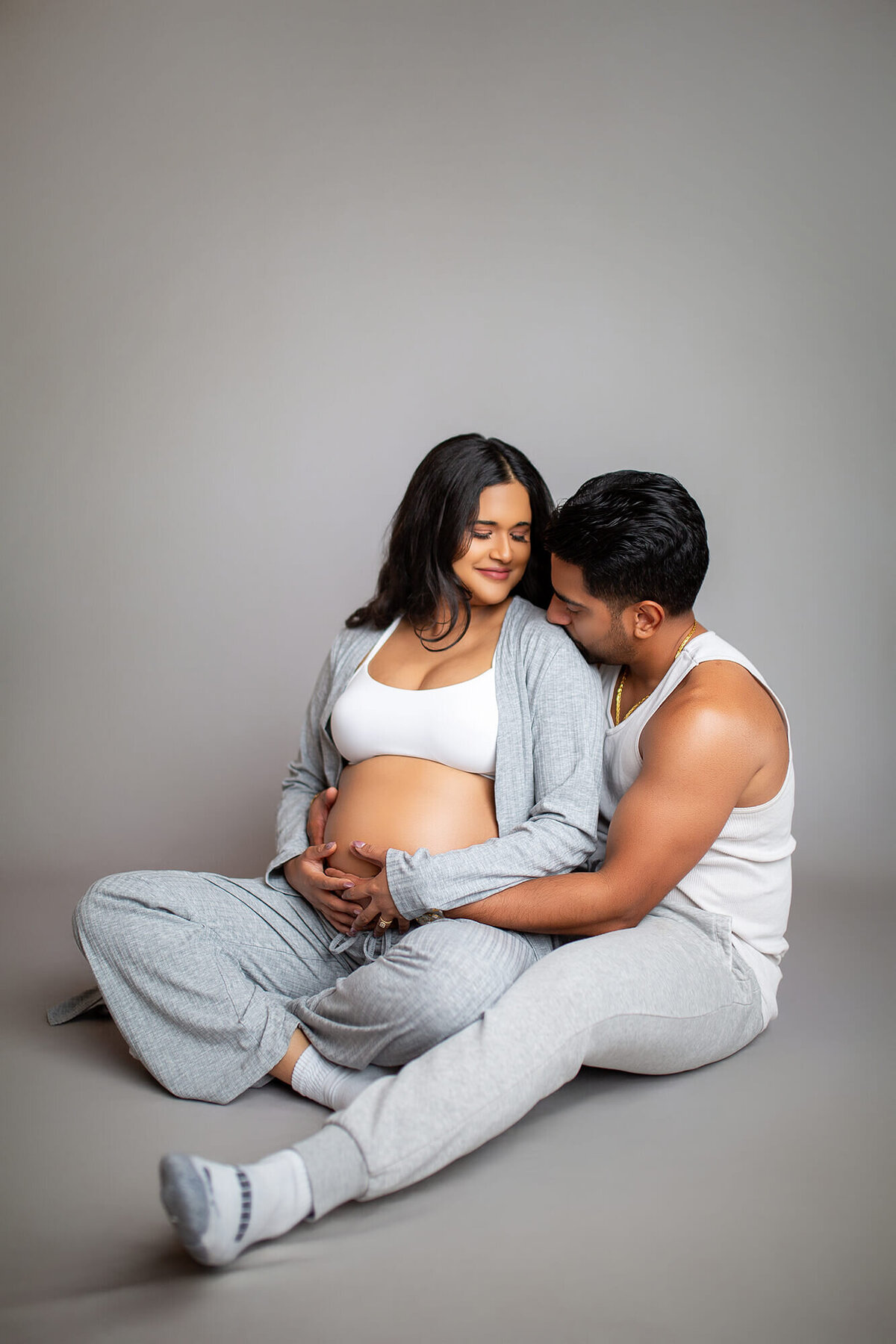 Cozy couple maternity studio session. Husband is kissing moms shoulder with arms wrapped around her. Studio located in Tampa, FL
