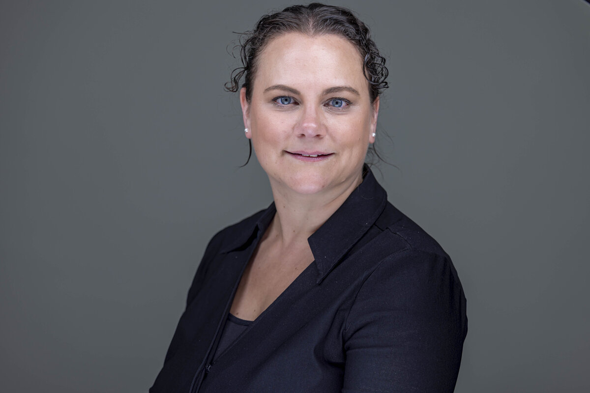 Professional headshot of a woman in a dark blazer, arms straight, smiling warmly in front of a grey flat background, by Rob Faber Photography, a headshot photographer in CT.