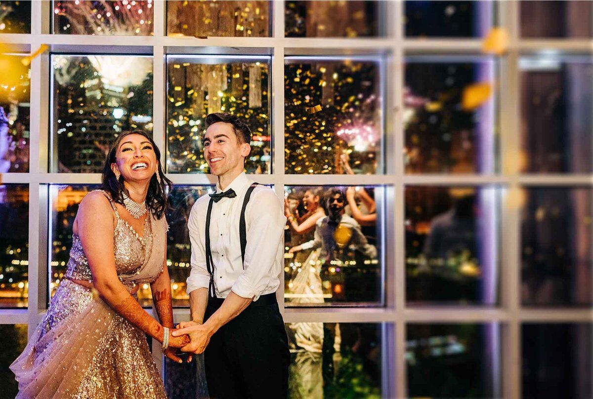 Bride in a sparkling gown and groom in suspenders laughing in front of a large window as fireworks and confetti light up the night sky.